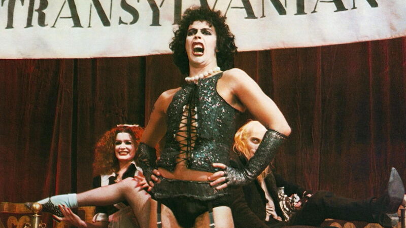 Tim Curry sings on stage while dress in a corset and suspenders