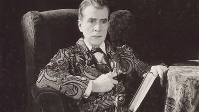 A black-and-white still shows a man in a patterned dressing gown smoking a pipe and sitting on an armchair.