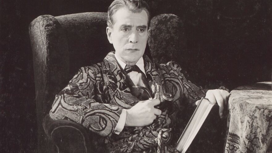 A black-and-white still shows a man in a patterned dressing gown smoking a pipe and sitting on an armchair.