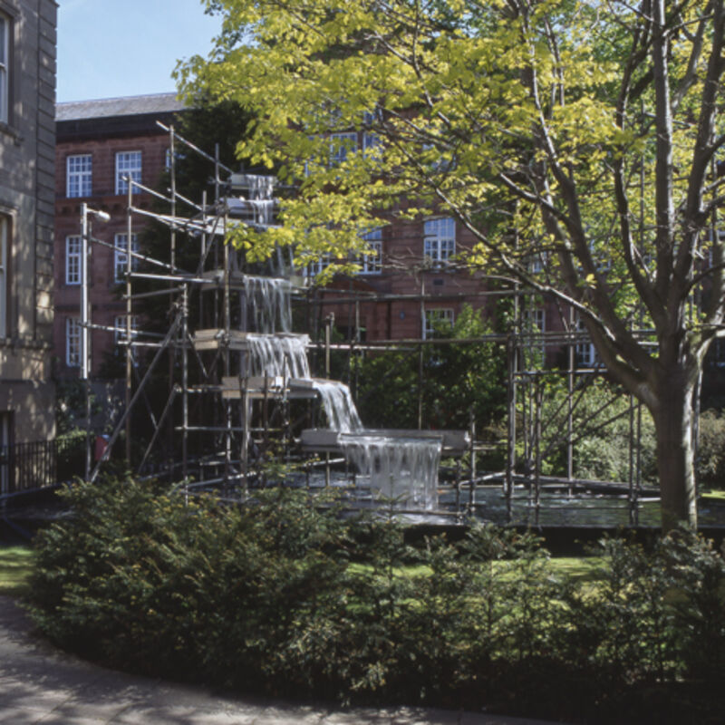 From Our Surroundings at DCA. A water feature made out of scaffolding sits in a garden with a tree in it. There is a 3-story high red building in the background.