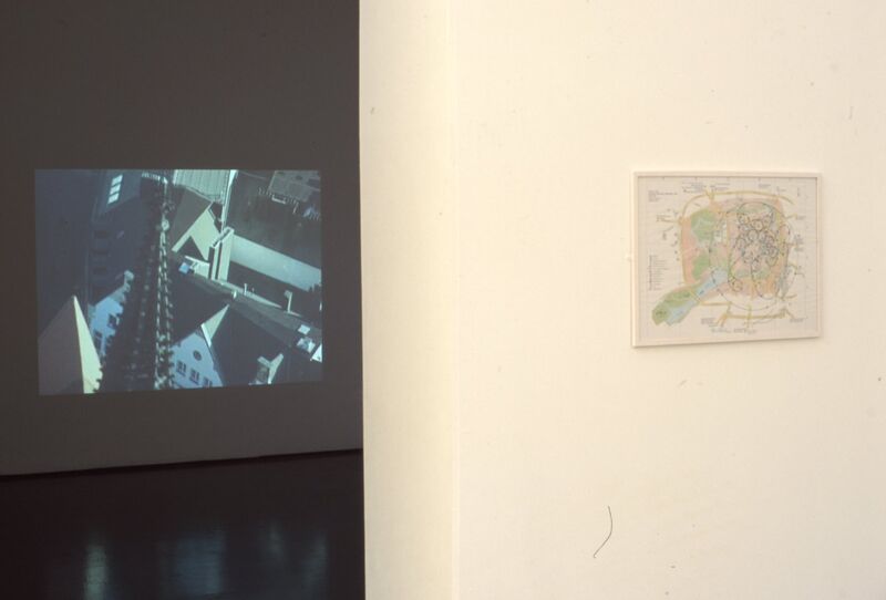 A map of a city hangs on a white wall. In the background, a projected image shows a birds-eye view of a city.