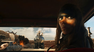 A woman with dark face paint on the top-half of her face sits in a car. Out of the car window, people are jumping on top of a truck, and there is a small explosion.