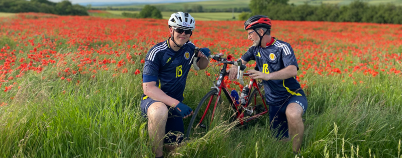 Two men wearing Scotland football strips pose in a poppy field holding a bike.