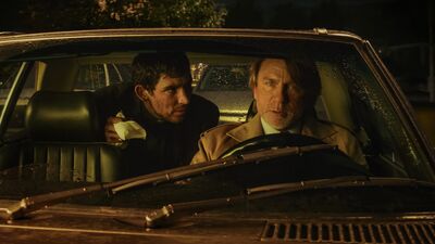 A worried man drives a vintage car while another man in the back seat leans over watching him