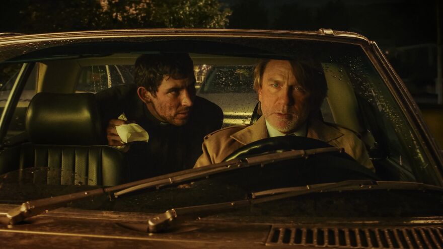 A worried man drives a vintage car while another man in the back seat leans over watching him