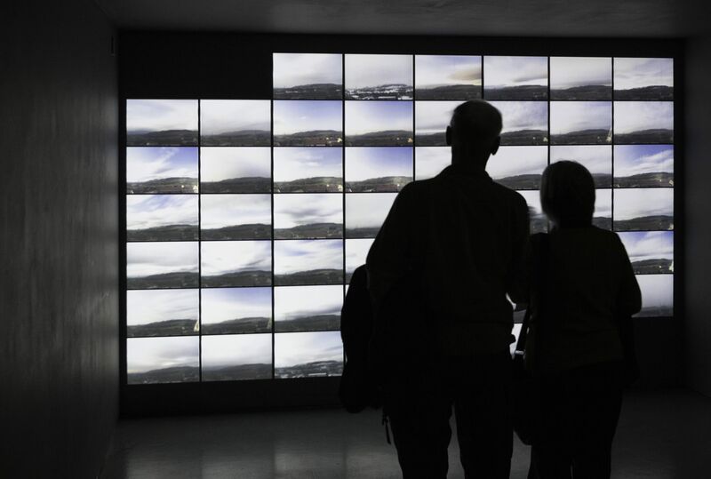 From Thomas & Craighead's exhibition. Two visitors to DCA Galleries look at multiple TV screens which show slightly different image of mountains and clouds.