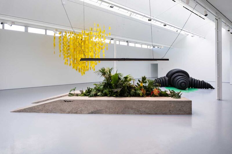 A gallery space: a car ramp with plants inside it and lights hanging above it in the background a yellow mobile and Michelin man in the backgroud
