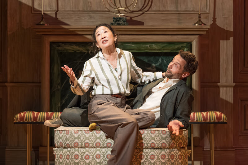 Two people sit on a sofa with their arms raised in a questioning shrug
