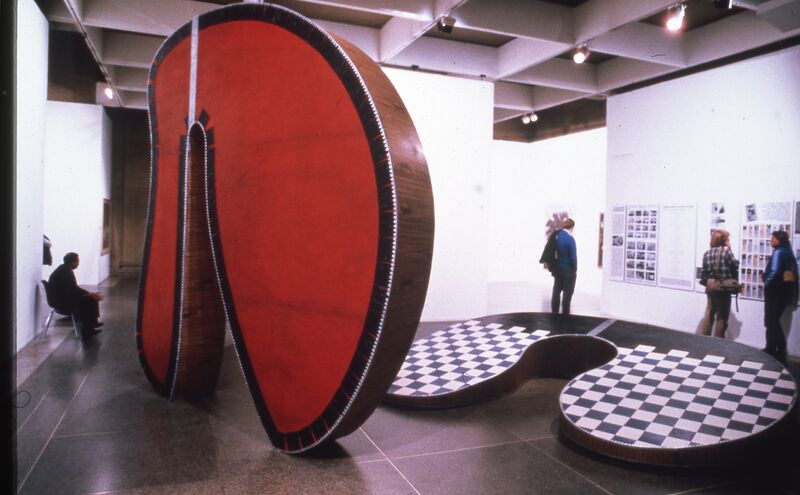 A large red, wooden statue shaped like a circle stands in DCA Galleries, next to a flat statue with a checkerboard pattern on it.
