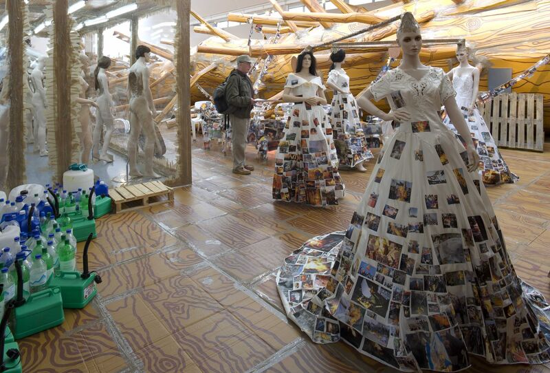 From Thomas Hirschhorn's exhibition. Mannequins are dressed in wedding dresses which are covered in pictures of burning buildings. There are plastic bottles in the corner of he room, and a large tree.