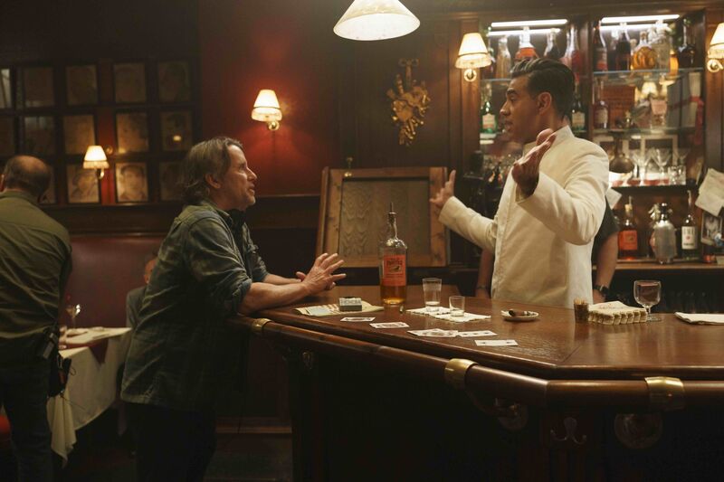 A man talks in a bar to a bartender wearing a white suit. A bottle of whisky and a pack of cards sit on the table