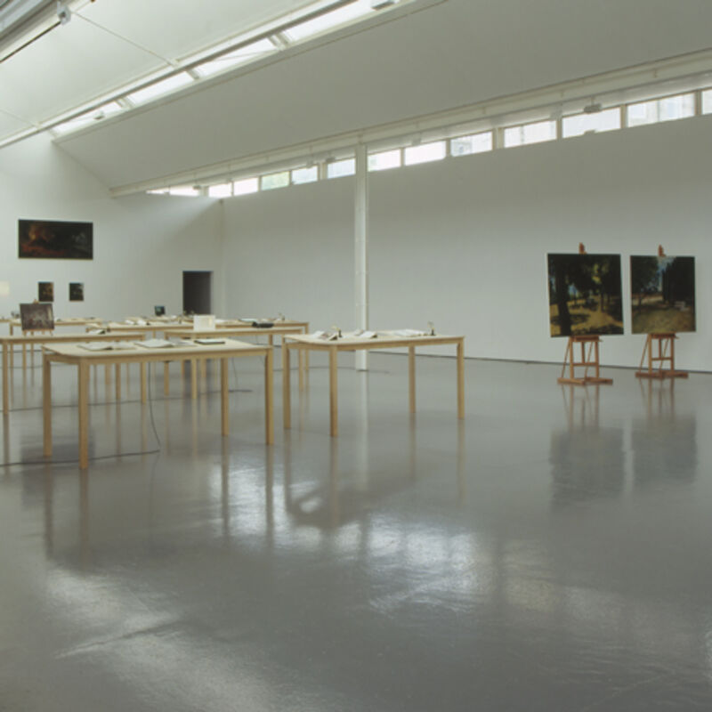 From Matts Leiderstam's exhibition in DCA Galleries. Tall tables stand in the galleries with books on them. To the right, two easles stand with landscape paintings on them.