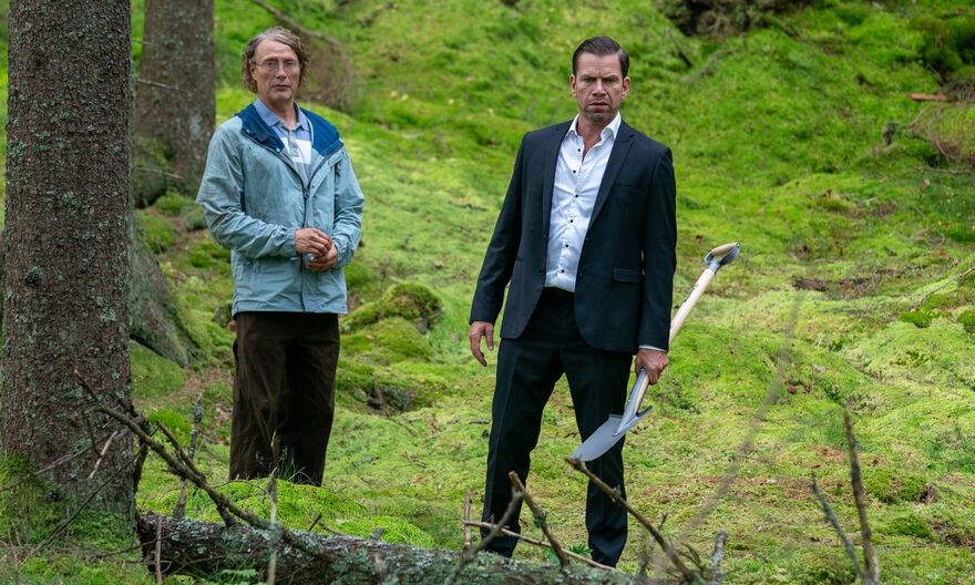 Two people stand in a moss-covered forest, with one person in a dark suit holding a shovel while the other wears an outdoor jacket, surrounded by trees and green woodland ground cover.