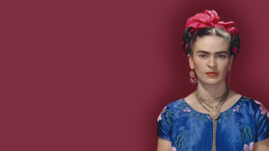 A photograph of Frida Kahlo wearing a blue dress, gold jewlerry and a red ribbon in her hair.