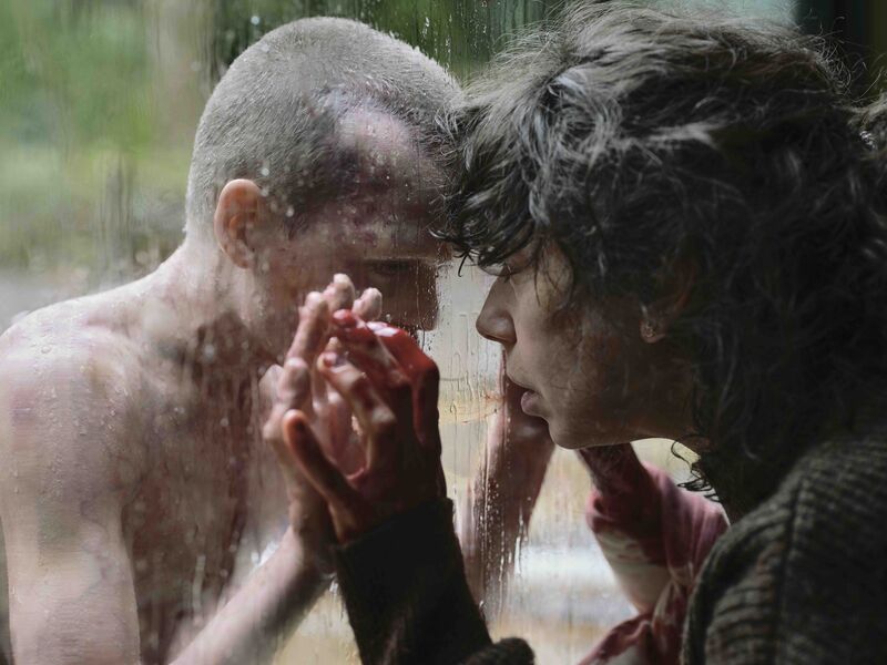 A woman looks at a child with a bald head through a window.