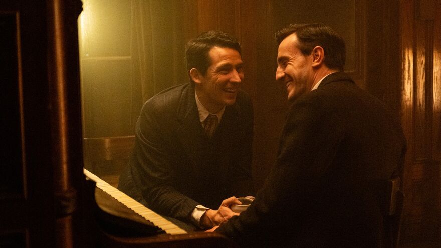Two dark-haired men in 1940s clothing laugh together at a piano