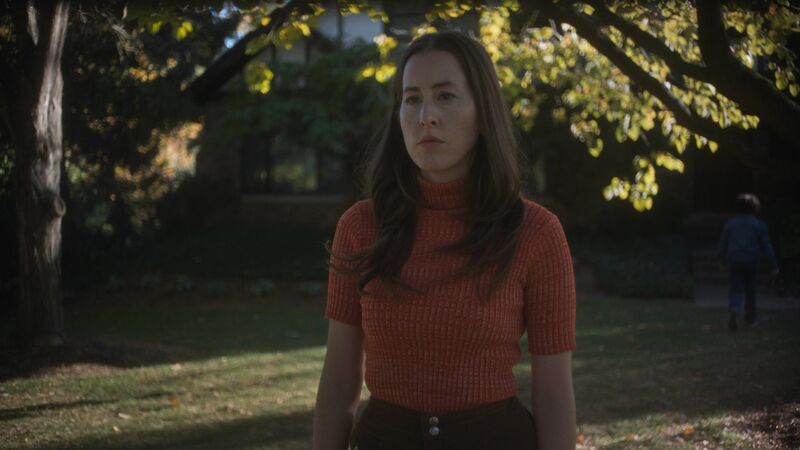 A woman wearing a red polo neck stands outside next to a tree