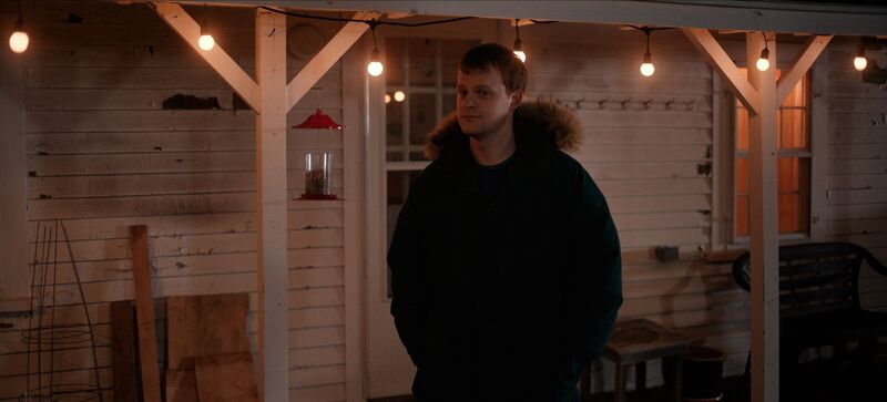 A young man stands in front of a porch hung with fairy lights.