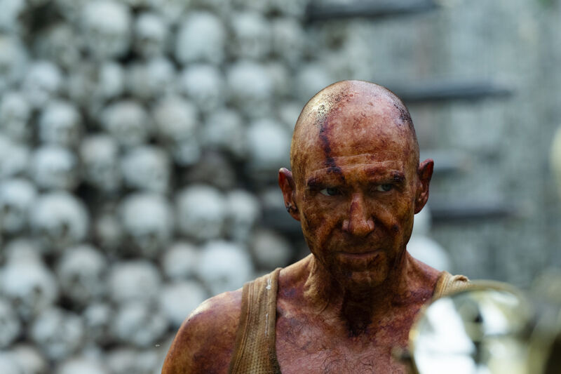 A man covered in dirt and blood stands next to a tower of skulls.