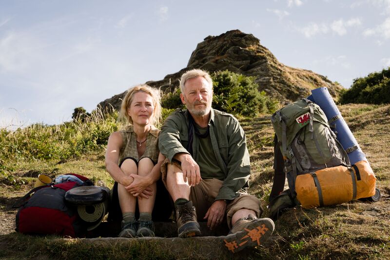 A middle-aged man and woman sit on the grass by a hill with hiking gear next to them.