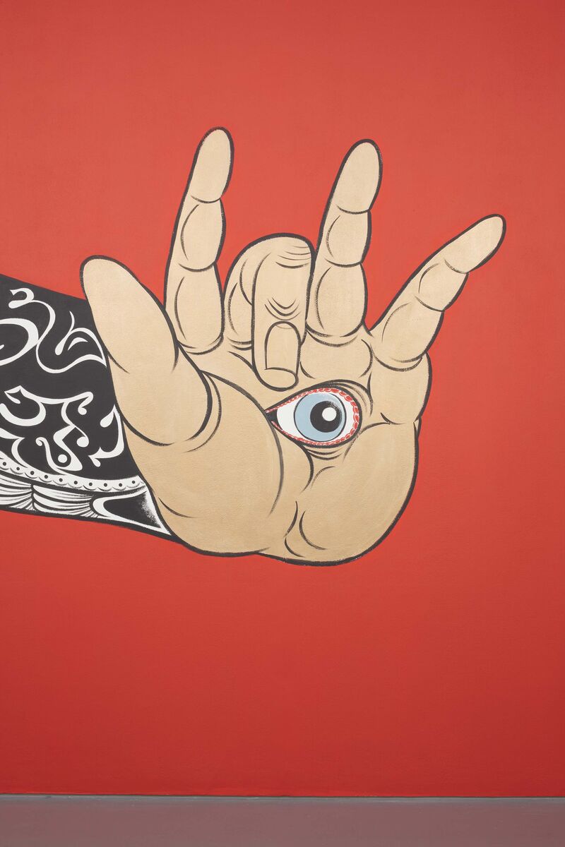 From Hideyuki Katsumata's exhibition - a mural shows a tattooed arm with a hand making the horn symbol. The hand has an eye on its palm. The background of the wall is red.