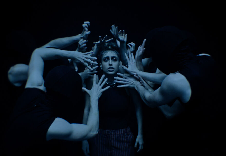 A young woman stands in the middle of a group of dancers with their hands reaching for her face