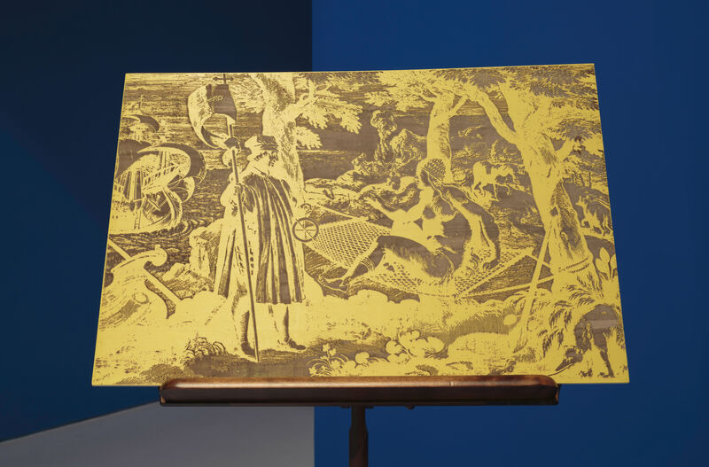 A photograph of an image displayed on a wooden stand. Bright blue walls can be seen in the background. The image shows a colonial era print, mainly in yellow and Cooper brown.  