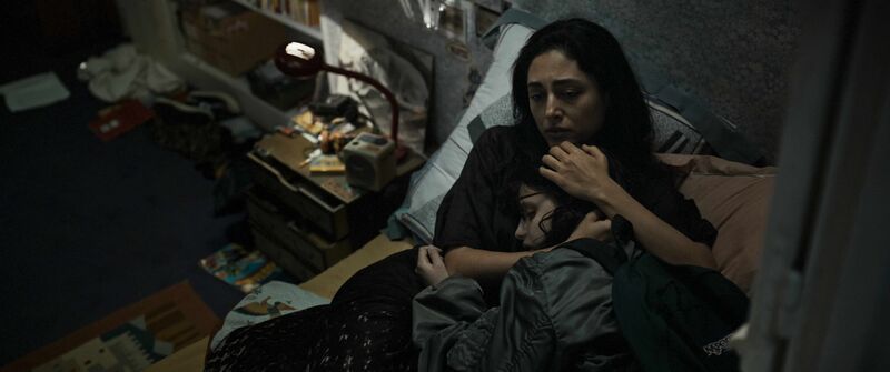 A mother lies on a bed, hugging her child and looking worried.