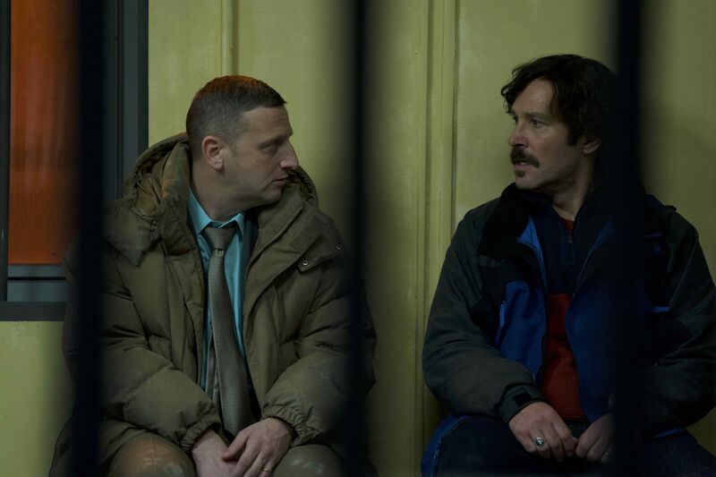 Two men, both wearing business clothes and jackets, look at each other with serious expressions.