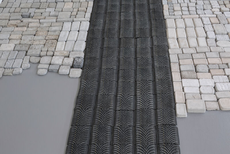 A series of cast bricks is shown laid out on the floor in a grid, either side of a long black strip of rubber in the centre of the image, made from car tyre. 