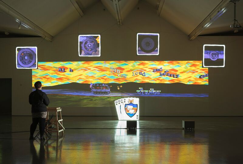 Eddo Stern's exhibition in DCA Galleries. Two large projectors show a mixed media image of a tank driving on a dirt track. The sky is made up of yellow, orange and green diamonds, and there are floating letters in the sky. An illuminated sculpture of a deck of cards is in front of the projector.