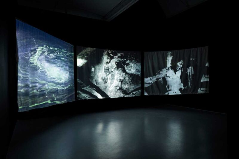 3 screens hanging on a wall - they have black and white images on them including a whirling water, horses galloping and a lady singing