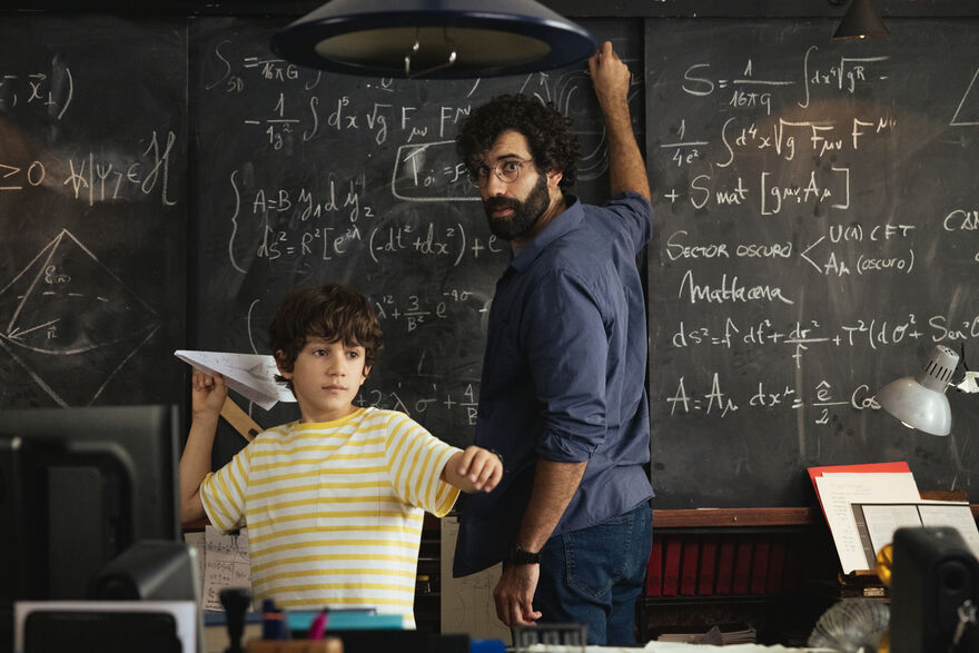 A bearded man looks at camera while writing on the blackboard and a small boy throws a paper aeroplane