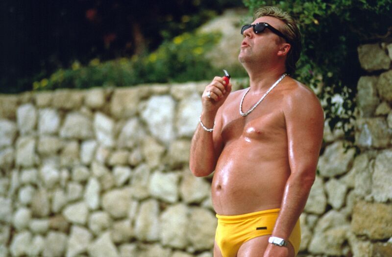 Ray Winstone smokes while wearing yellow swim trunks