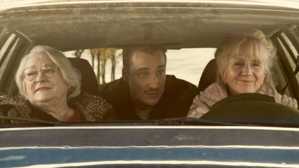 Two women sit in a car, smiling. A man sits in the back seat talking to them.