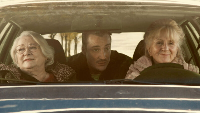 Two women sit in a car, smiling. A man sits in the back seat talking to them.