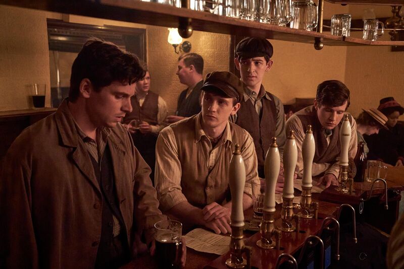 A group of young men in 1910s attire sit in a crowded bar together