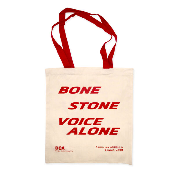 Cream tote bag with red text reading BONE STONE VOICE ALONE and red handles. Printed smaller at the bottom of the bag is red DCA logo and text: A major new exhibition by Lauren Gault