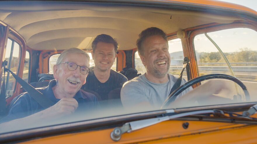 Two men and an elderly man drive an orange car together, laughing.