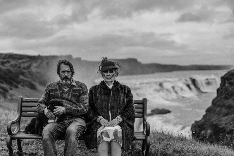 A black and white image of a man and a old woman sitting on a bench, in front of dramatic cliffs and ocean.