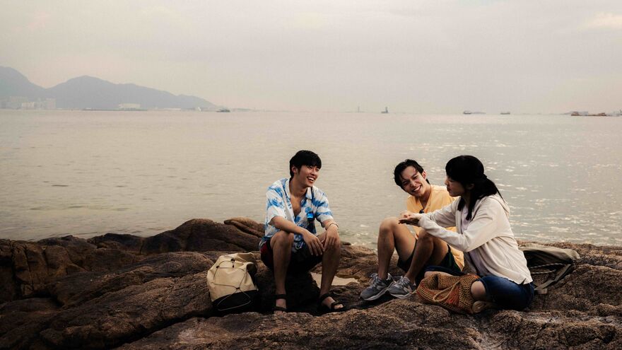 Three people sit on rocks at a beach together, talking to each other in sign language