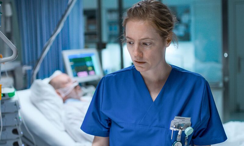 A nurse in a blue uniform stands in front of a hospital bed looking nervous