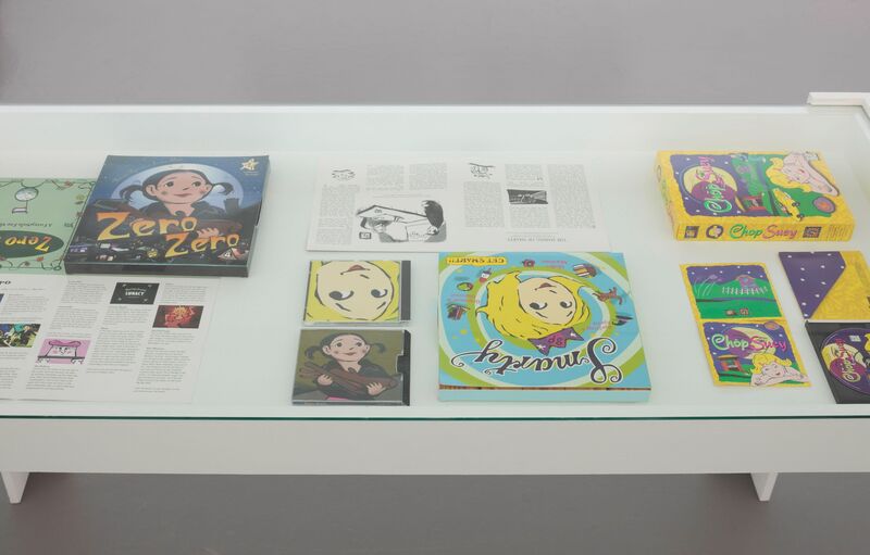 A gladd table display at Theresa Duncan's exhibition shows miscellaneous items from the production of her three games, including CD and video tape covers of Sunday, Chop Suey and Zero Zero. Each cover features a computer drawing of a young girl.