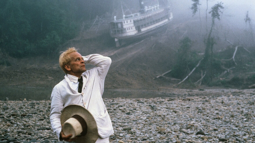 A man in a white suit stands on an abandoned beach with a wrecked ship in the background