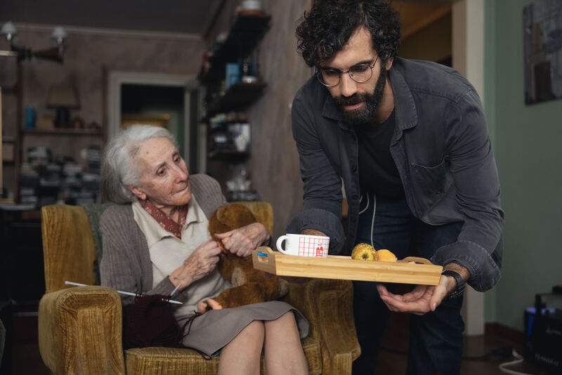 A man is giving an elderly lady a tray with a mug and fruit