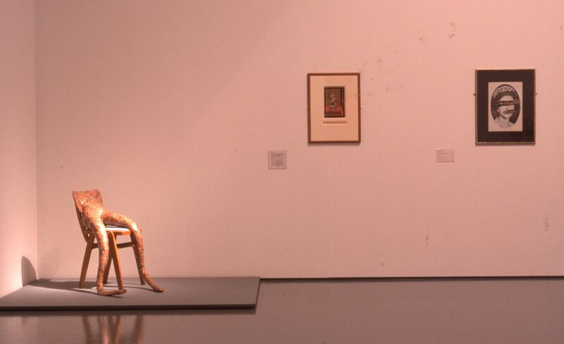 A sculpture of legs sitting on a chair sits in DCA Galleries, next to two small framed portraits.