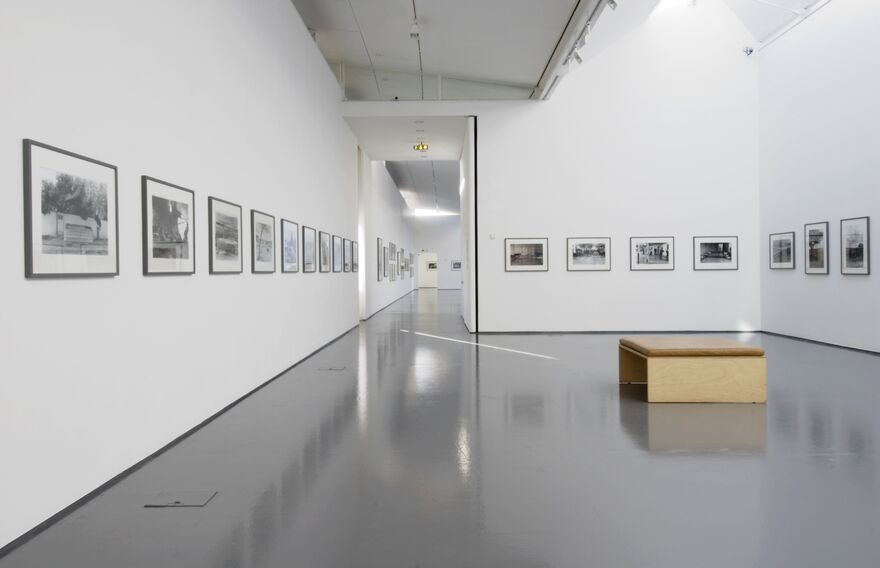 DCA Galleries during Ahlam Shibli's exhibition. The gallery is brightly lit, and medium-sized black-and-white photographs are on the walls. There is a bench in the middle of the room. 