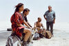 Three men and a woman sit with luggage in a barren desert.