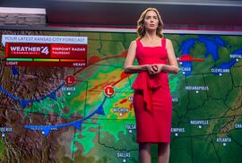 A woman in a red dress stands in front of a digital screen with a weather map behind her