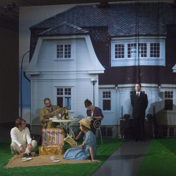 From Killing Time at DCA Galleries. Four people have a picnic on fake gras in the galleries in front of a large fabric sheet with a photograph of a house on it. There is a real wooden shed next to the people.